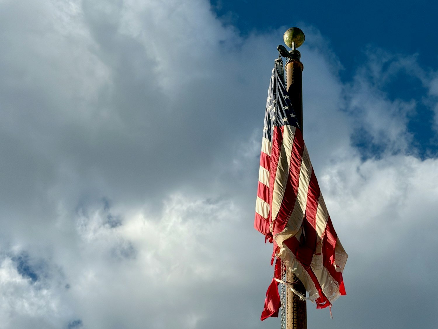 A tattered American flag waving against a cloudy sky, showing the wear that signals it should be retired